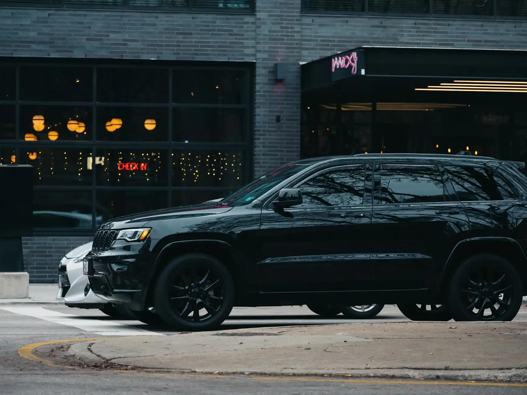 premium black town car and full-size SUV side by side outside Sea-Tac arrivals area at dusk, travelers loading suitcases, terminal lights reflecting on wet pavement