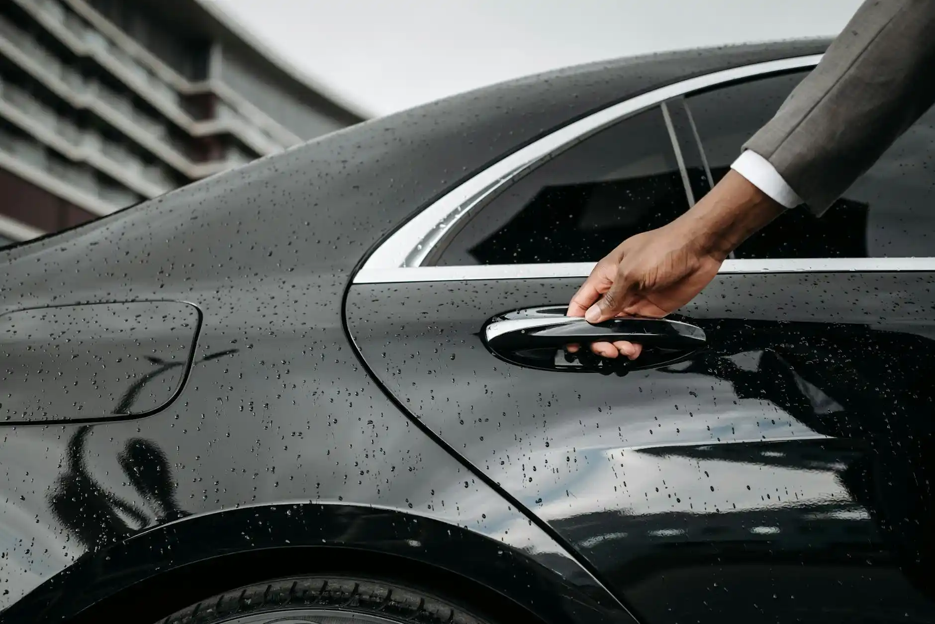 luxury black town car parked in designated airport pickup zone at Sea-Tac, traveler exiting terminal with rolling luggage under covered curbside area, wet pavement reflecting ci...