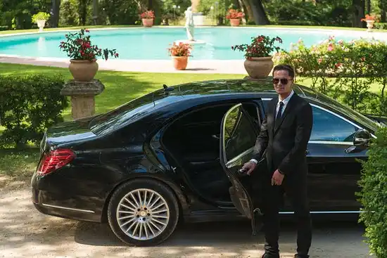 professional chauffeur opening rear door of a dark executive sedan outside a downtown Seattle office tower, early morning light, traveler in business attire holding briefcase