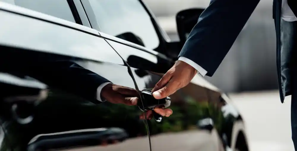 Uniformed chauffeur checking a black executive van before a SeaTac transfer, inspecting tires and luggage area with Seattle skyline faintly visible